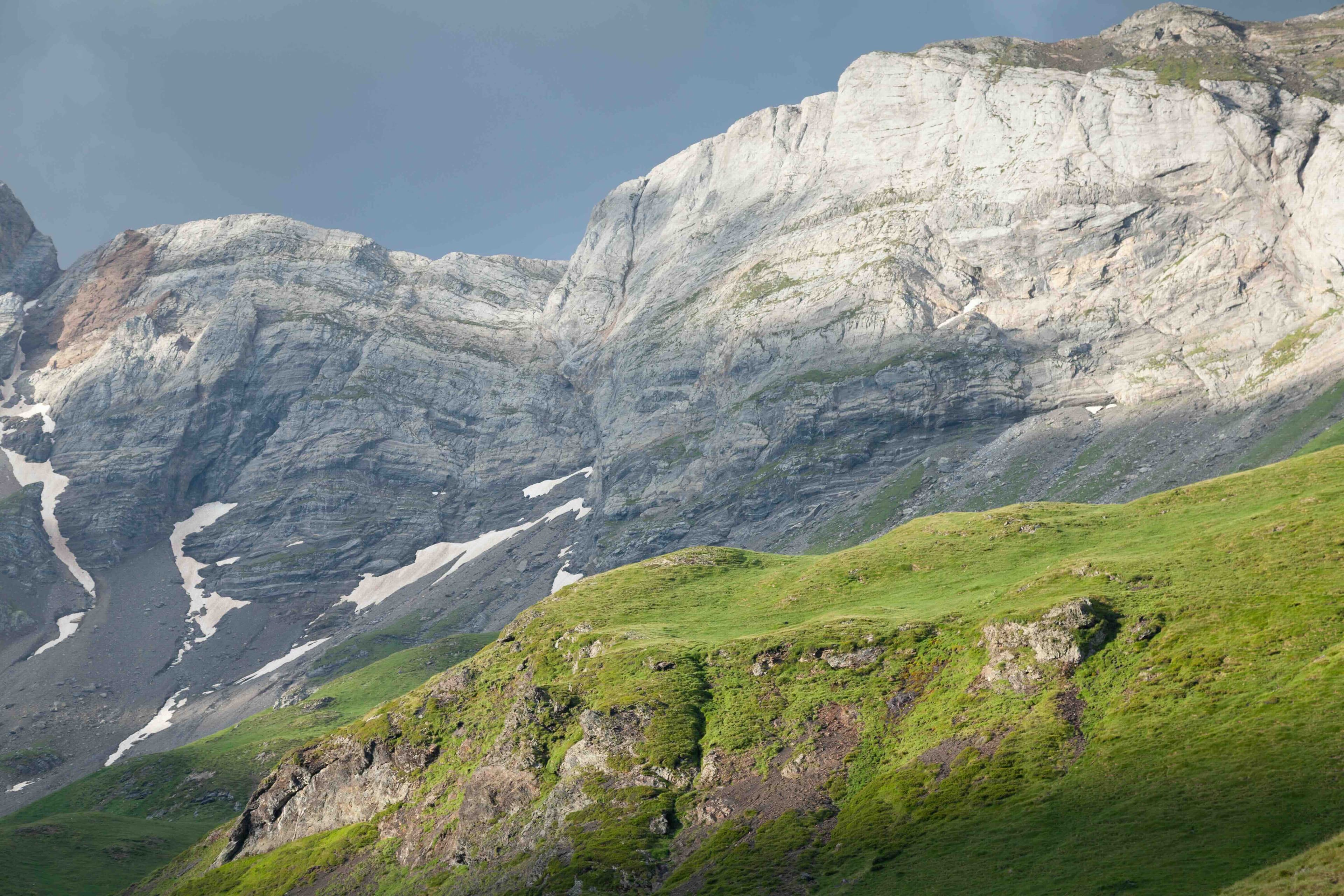 Steile Kalkfelswand mit Schneeresten über grünen Almwiesen im Hochgebirge, dunkler Gewitterhimmel. Symbolbild für alpine Destinationen.